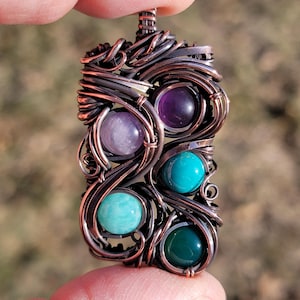 Peut inclure: Pendentif artisanal de forme rectangulaire, réalisé avec du fil de cuivre. Il est orné de cinq pierres précieuses polies de couleurs violet, turquoise et vert. Les pierres sont intégrées dans le fil, créant un bijou unique.