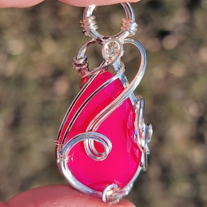 May include: A handcrafted pendant featuring a vibrant, teardrop-shaped, hot pink gemstone. The stone is encased in intricate, swirling silver wire, with a looped bail for a chain. The pendant is held between fingers against a blurred natural background.