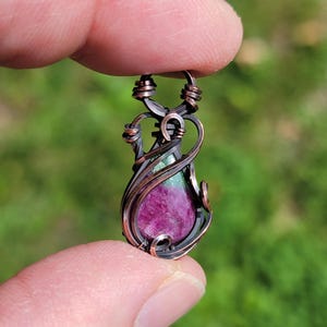 May include: A handcrafted copper pendant featuring a teardrop-shaped ruby gemstone. The ruby is a deep magenta color, accented by green inclusions. The pendant is wire-wrapped in a decorative pattern.