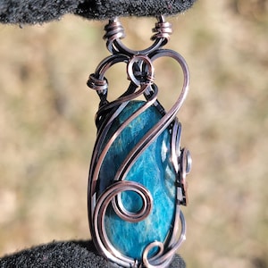 May include: A handcrafted pendant featuring a teardrop-shaped, vibrant teal stone. The stone is encased in intricate wirework with copper and black metal accents. The pendant is suspended from a matching wire loop, showcasing a unique, artistic design.