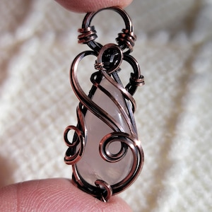 May include: A handcrafted pendant featuring a teardrop-shaped, pale pink stone encased in copper wire. The wire is intricately wrapped and curled, creating an ornate, artistic design. The pendant is held by a finger.