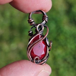 May include: A teardrop-shaped red gemstone pendant encased in dark, oxidized copper wire wrapping. The wire forms intricate swirls and loops around the faceted stone, culminating in a circular bail at the top for hanging on a chain or cord.