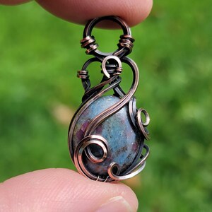 May include: A wire-wrapped pendant featuring a smooth, teardrop-shaped blue and magenta stone encased in dark, oxidised copper wire. The metalwork consists of intricate swirls and coils that form a decorative frame around the gemstone.