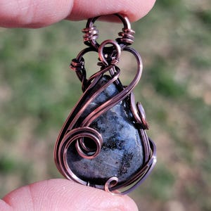 May include: A handcrafted pendant featuring a teardrop-shaped, dark blue gemstone with black inclusions, intricately wrapped in copper wire. The wirework includes decorative swirls and loops, creating a unique, artistic design. The pendant is held up by a person's fingers.
