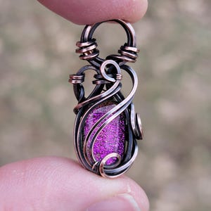 May include: A handcrafted pendant featuring a vibrant purple dichroic glass stone encased in intricate copper wire. The wirework is a mix of dark and rose-gold tones, creating a unique, artistic design. The pendant is held up by a finger.