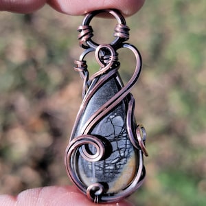 May include: A handcrafted pendant featuring a teardrop-shaped stone with a unique gray and tan pattern. The stone is intricately wrapped in copper wire, creating a decorative frame and bail. The pendant is held between fingers.