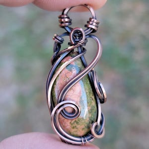 May include: A handcrafted pendant featuring a teardrop-shaped stone with green and pink hues, encased in a copper wire wrap. The wire is intricately designed with swirls and loops, creating a unique and artistic jewelry piece. The pendant is held up by a finger.