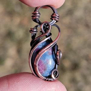May include: A handcrafted pendant featuring a blue and pink gemstone, intricately wrapped in copper and dark metal wire. The teardrop-shaped stone is the focal point, with the wire forming decorative swirls and a loop for a chain. The pendant is held between fingers.