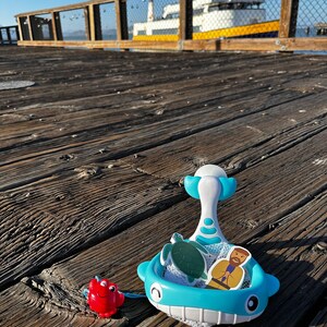 May include: A blue whale-shaped toy with a net and a white handle. The toy is filled with a green turtle and a yellow figure of a person. A red crab toy is on the wooden surface.