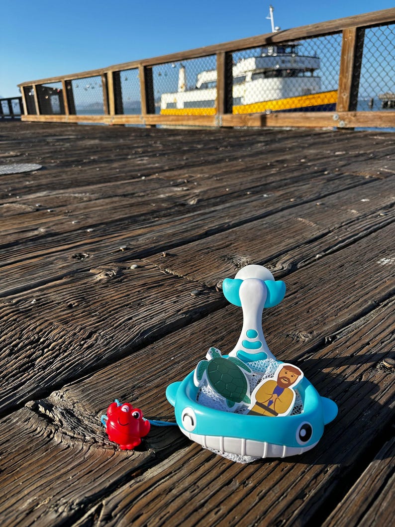 May include: A blue whale-shaped toy with a net and a white handle. The toy is filled with a green turtle and a yellow figure of a person. A red crab toy is on the wooden surface.