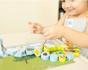 May include: A child plays with a wooden toy featuring a colorful string of beads with images of people and objects. The beads are strung on a red string and the child is using their fingers to thread the string through the beads. The beads are on a blue and green background with clouds and trees.