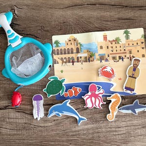 May include: A blue whale-shaped net with a white mesh and a red handle. The net is surrounded by various sea creatures, including a turtle, a jellyfish, an octopus, a dolphin, a clownfish, a shark, and a seahorse. The background is a colorful illustration of a seaside town.