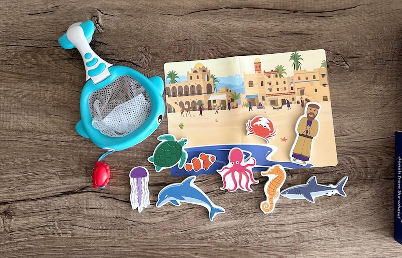 May include: A blue whale-shaped net with a white mesh and a red handle. The net is surrounded by various sea creatures, including a turtle, a jellyfish, an octopus, a dolphin, a clownfish, a shark, and a seahorse. The background is a colorful illustration of a seaside town.