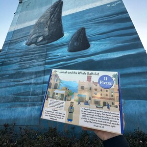 May include: A colorful box for a bath toy set featuring Jonah and the Whale. The box has a picture of a city with a blue sky and a whale in the water. The text on the box reads "3yrt Jonah and the Whale Bath Set 11 Pieces". The text on the back of the box reads "So they took up Jonah and threw him into the sea, and the sea ceased from its raging. Then the men feared the LORD exceedingly, and they offered a sacrifice to the LORD and made vows. And the LORD appointed a great fish to swallow up Jonah, and Jonah was in the belly of the fish three days and three nights. Jonah 1:15-17"