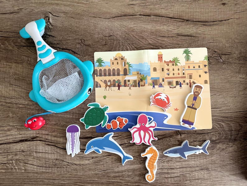 May include: A blue plastic toy net with a handle, a red crab, a green turtle, a purple jellyfish, a blue dolphin, a red and orange fish, a pink octopus, a yellow seahorse, and a gray shark. The toys are on a wooden surface with a background of a colorful illustration of a city.
