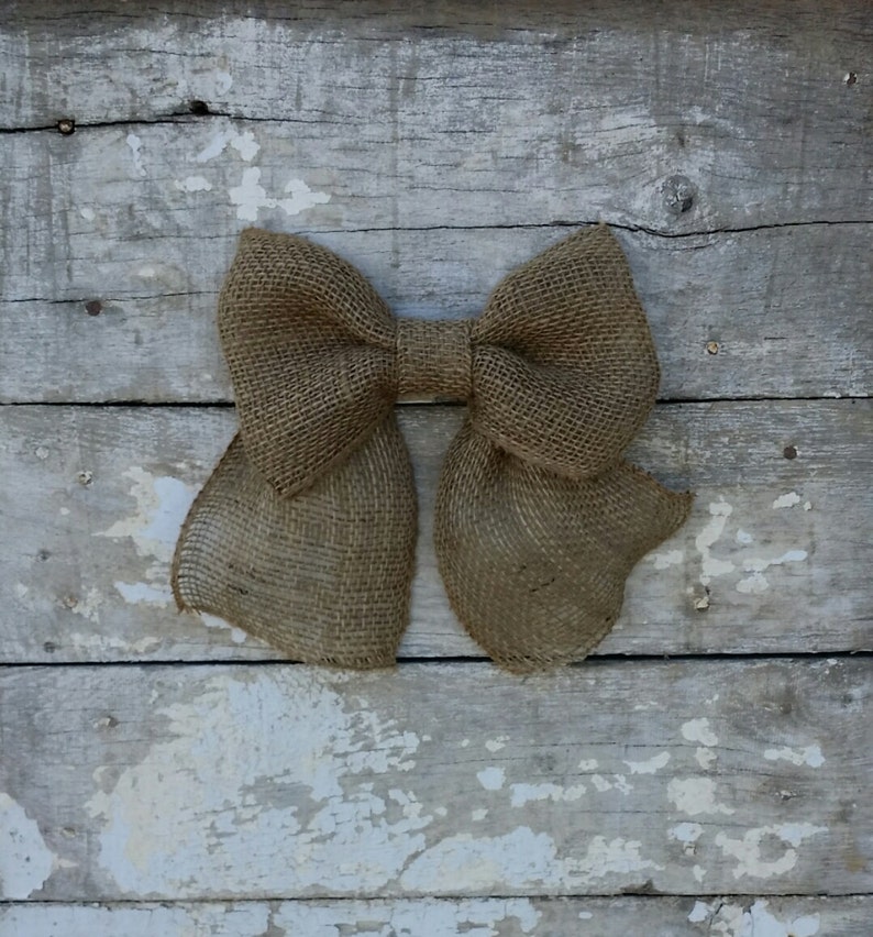 May include: A large, rustic burlap bow with a classic bow shape. The bow is a natural tan color and is set against a weathered, white-painted wooden background. The bow's texture is coarse and woven.