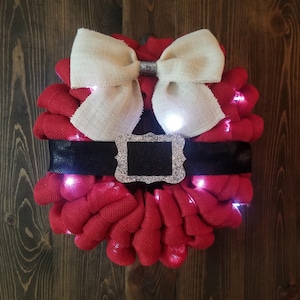 May include: A festive Christmas wreath with a Santa Claus theme. The wreath features a red burlap base, a black belt with a silver buckle, and a cream-colored burlap bow. Small white lights are interspersed throughout the wreath.