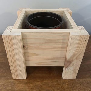 May include: A natural wood planter box with a dark brown pot inside. The planter box is made of light-colored wood and has a rectangular shape with four legs. The pot is visible inside the box, ready for planting.