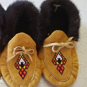 Könnte beinhalten: Hellbraune Wildleder-Mokassins mit einem geometrischen Perlenmuster in Rot, Gelb, Schwarz und Weiß. Die Oberseite der Mokassins ist mit dunkelbraunem Fell gefüttert. Traditioneller Stil.