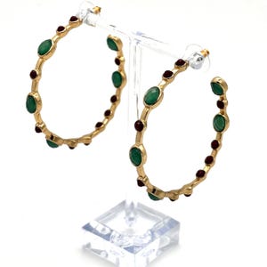 May include: A pair of gold hoop earrings featuring alternating round green and dark red stones set along the circular metal bands. The earrings are displayed on a clear plastic stand with post-style backings visible at the top of each hoop.