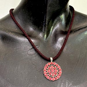 May include: A necklace featuring a dark brown braided cord and a circular silver-toned pendant. The pendant is decorated with a radial pattern of small, teardrop-shaped coral-colored stones arranged in concentric circles around a central stone.