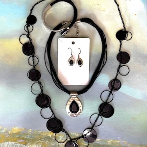 May include: A black jewelry set featuring a necklace with circular and disc-shaped accents, a matching bracelet, and teardrop earrings. The necklace has a pendant with a black stone. The jewelry is displayed on a white card.