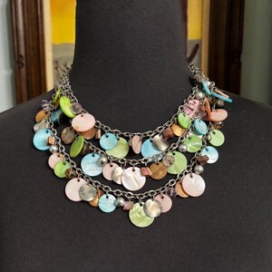 May include: A multi-strand necklace with a silver chain and colorful, iridescent, circular pendants. The pendants are in shades of pink, blue, green, and brown. The necklace is displayed on a black mannequin.