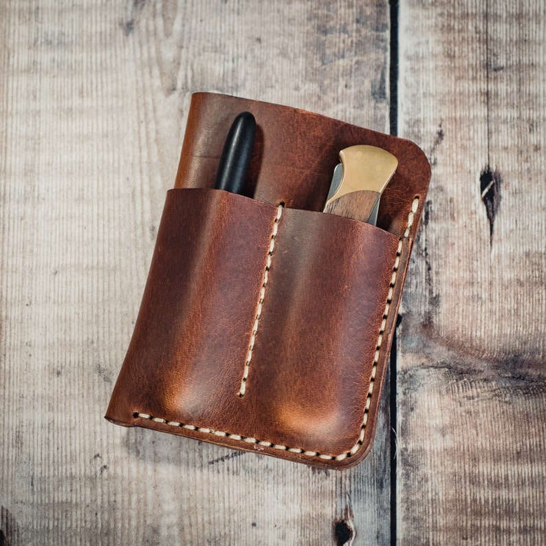 May include: A brown leather pocket organizer with two slots, one for a pen and one for a small knife. The organizer has a stitched edge and is made of distressed leather.