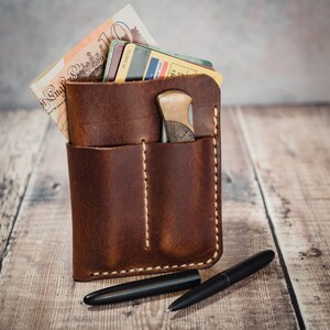 May include: A brown leather wallet with a stitched pocket and a small knife inside. The wallet is sitting on a wooden surface with a black pen next to it.
