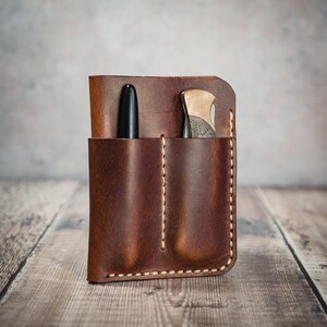 May include: A brown leather pocket organizer with two pockets, one for a pen and one for a small knife. The organizer has a stitched edge and is made of distressed leather.