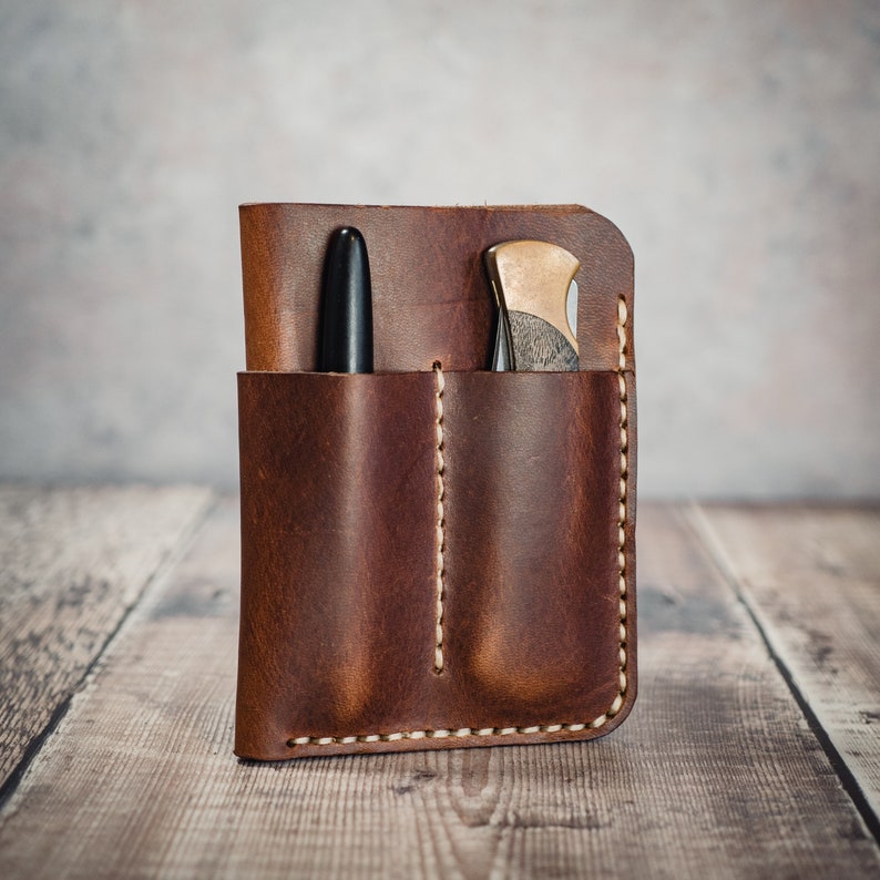May include: A brown leather pocket organizer with two pockets, one for a pen and one for a small knife. The organizer has a stitched edge and is made of distressed leather.