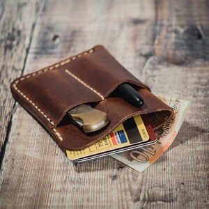 May include: A brown leather wallet with two pockets, a pen, and a small knife inside. The wallet is open and the contents are visible. The wallet is sitting on a wooden surface.