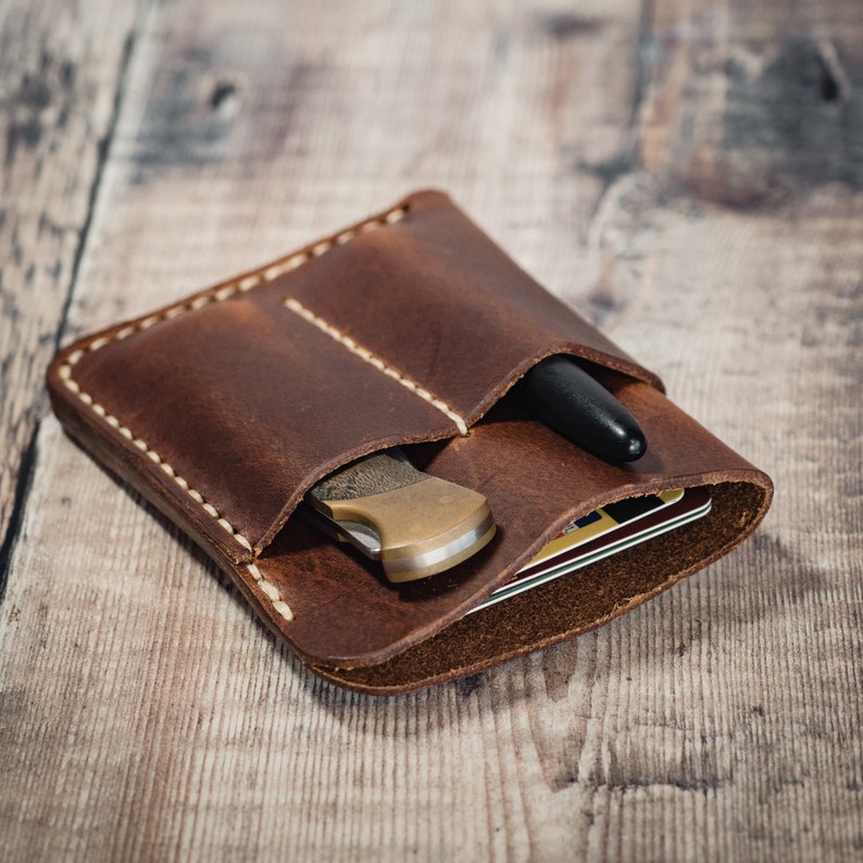 May include: A brown leather wallet with two pockets, one for a pen and one for a small knife. The wallet is lying on a wooden surface.