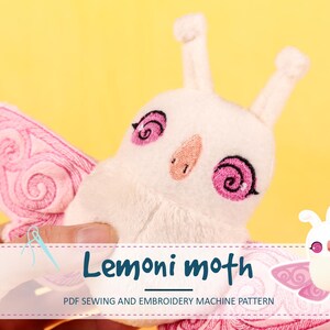 Könnte beinhalten: Ein weißes Plüsch-Motten-Spielzeug mit rosa gestickten Details, einschließlich wirbelnder Augen und Flügelmustern. Die Motte hat eine hellrosa Nase und Fühler. Der Text "Lemoni moth" und "PDF Sewing and Embroidery Machine Pattern" ist enthalten.
