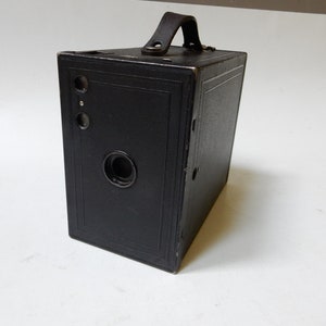 May include: A vintage black box camera with a leather handle. The camera has a rectangular shape with a lens and two viewfinders on the front. The camera is made of a dark material and has a handle on top.