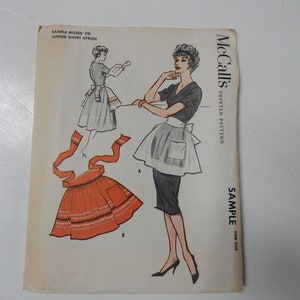 May include: Vintage McCall's sewing pattern for a misses' or junior short apron. The pattern is printed on a white paper with a black and white illustration of a woman wearing the apron. The text "SAMPLE" and "ONE SIZE" is printed on the right side of the paper.