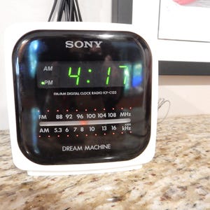 May include: A white Sony Dream Machine ICF-C122 digital clock radio with a black front panel. The display shows the time 4:17 in green digital numbers. The interface includes FM and AM radio frequency scales with a horizontal tuning indicator.