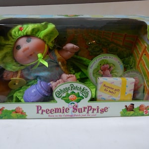 May include: A Cabbage Patch Kids Preemie Surprise doll in a green and blue outfit with a green hat. The doll is sitting in a green cabbage patch with a purple bottle and a pacifier. The box has the text "Surprise gift for baby!" and "Born in the Cabbage Patch, just for you!"