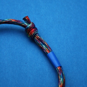 May include: A colourful rope bracelet with a blue knot and a blue stripe. The bracelet is on a blue background.