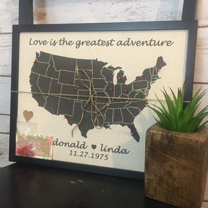 以下が含まれることがあります： アメリカ合衆国の地図、赤いピンマーカー、紐のディテールが施された額入りのキャンバスアート。上部には「Love is the greatest adventure」の文字、下部には名前と日付があります。右側には多肉植物が入った小さな木製のプランターがあります。