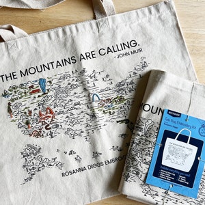 May include: Two beige tote bags with black handles. The larger bag has a black and white illustration of the United States with a map of national parks and the text "The Mountains Are Calling. -John Muir" and "Rosanna Diggs Embroidery". The smaller bag has the same illustration and text. A blue card with the text "Beginner Tote Bag Embroidery Kit" is in front of the smaller bag.