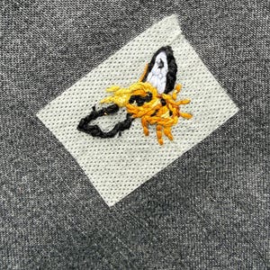 Pode incluir: Close-up de um tecido cinza com um desenho de abelha costurada. A abelha &eacute; bordada com fio preto, branco e amarelo em um patch cinza claro. O tecido tem uma apar&ecirc;ncia texturizada.