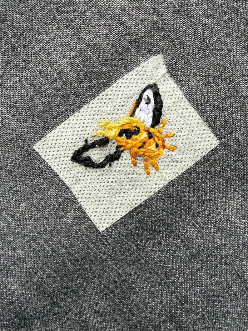 Pode incluir: Close-up de um tecido cinza com um desenho de abelha costurada. A abelha &eacute; bordada com fio preto, branco e amarelo em um patch cinza claro. O tecido tem uma apar&ecirc;ncia texturizada.
