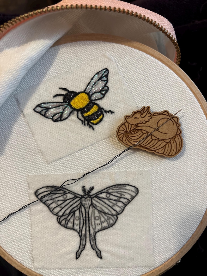 Pode incluir: Bastidor de bordado com tecido branco com uma abelha amarela e preta, uma mariposa e um drag&atilde;o de madeira. A abelha tem asas iridescentes. A mariposa &eacute; preta e branca. Um fio preto est&aacute; preso ao tecido.