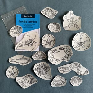 Pode incluir: Uma cole&ccedil;&atilde;o de tatuagens t&ecirc;xteis com v&aacute;rias criaturas marinhas e conchas, incluindo uma estrela do mar, um caranguejo e camar&otilde;es. As tatuagens s&atilde;o em arte de linha preta sobre tecido bege. Um pacote de "Beachcomber Textile Tattoos" tamb&eacute;m est&aacute; inclu&iacute;do.