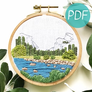 May include: Embroidered hoop art featuring a landscape scene with mountains, trees, a river, and rocks. The embroidery is done in shades of green, blue, and brown.