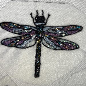 Pode incluir: Lib&eacute;lula bordada com asas iridescentes em tons de roxo, azul e prata, sobre um fundo branco texturizado. A lib&eacute;lula tem corpo e cabe&ccedil;a pretos, e as asas s&atilde;o detalhadas com detalhes brilhantes.