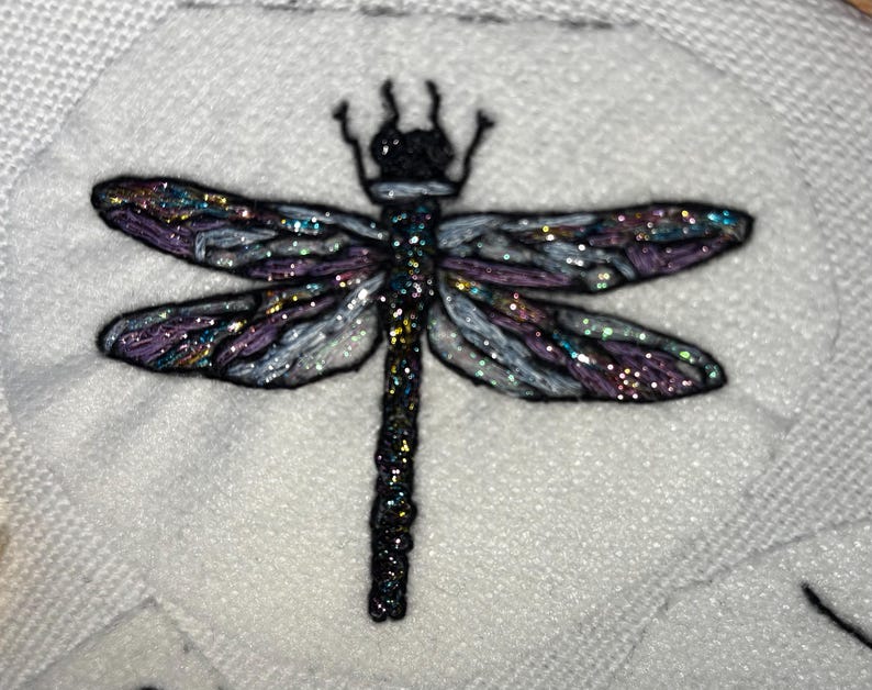 Pode incluir: Lib&eacute;lula bordada com asas iridescentes em tons de roxo, azul e prata, sobre um fundo branco texturizado. A lib&eacute;lula tem corpo e cabe&ccedil;a pretos, e as asas s&atilde;o detalhadas com detalhes brilhantes.