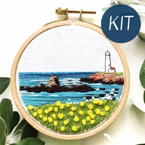 May include: A needlepoint embroidery kit featuring a coastal scene with a white lighthouse, blue water, and yellow wildflowers. The kit includes a hoop, fabric, and embroidery floss.