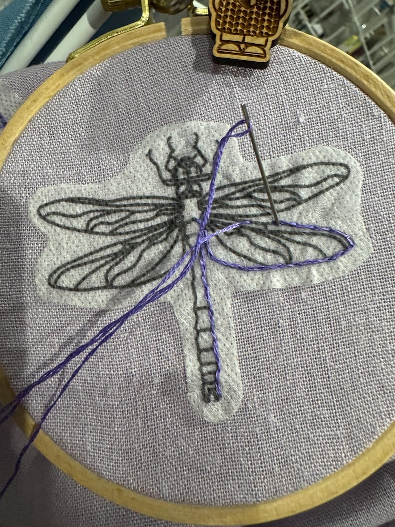Pode incluir: Um bastidor de bordado com tecido lavanda apresentando um desenho de lib&eacute;lula. A lib&eacute;lula &eacute; delineada em preto, com fio roxo usado para costurar o desenho. Uma agulha &eacute; inserida no tecido e uma presilha de madeira &eacute; presa ao bastidor.